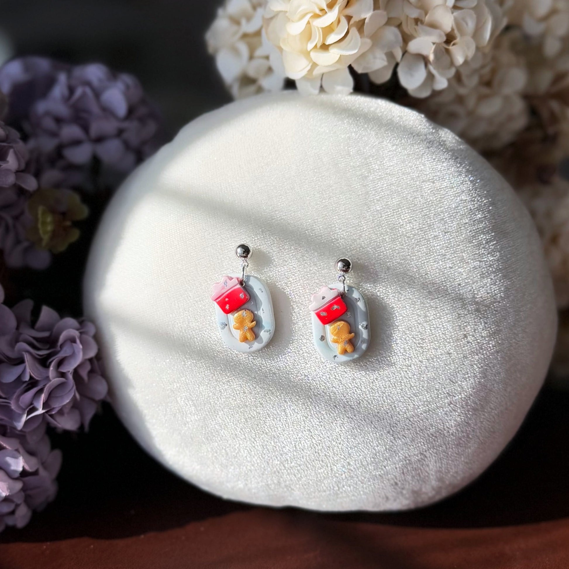 Pair of earrings with a Gingerbread Man and a gift box design on a white cushion surrounded by flower at the background.