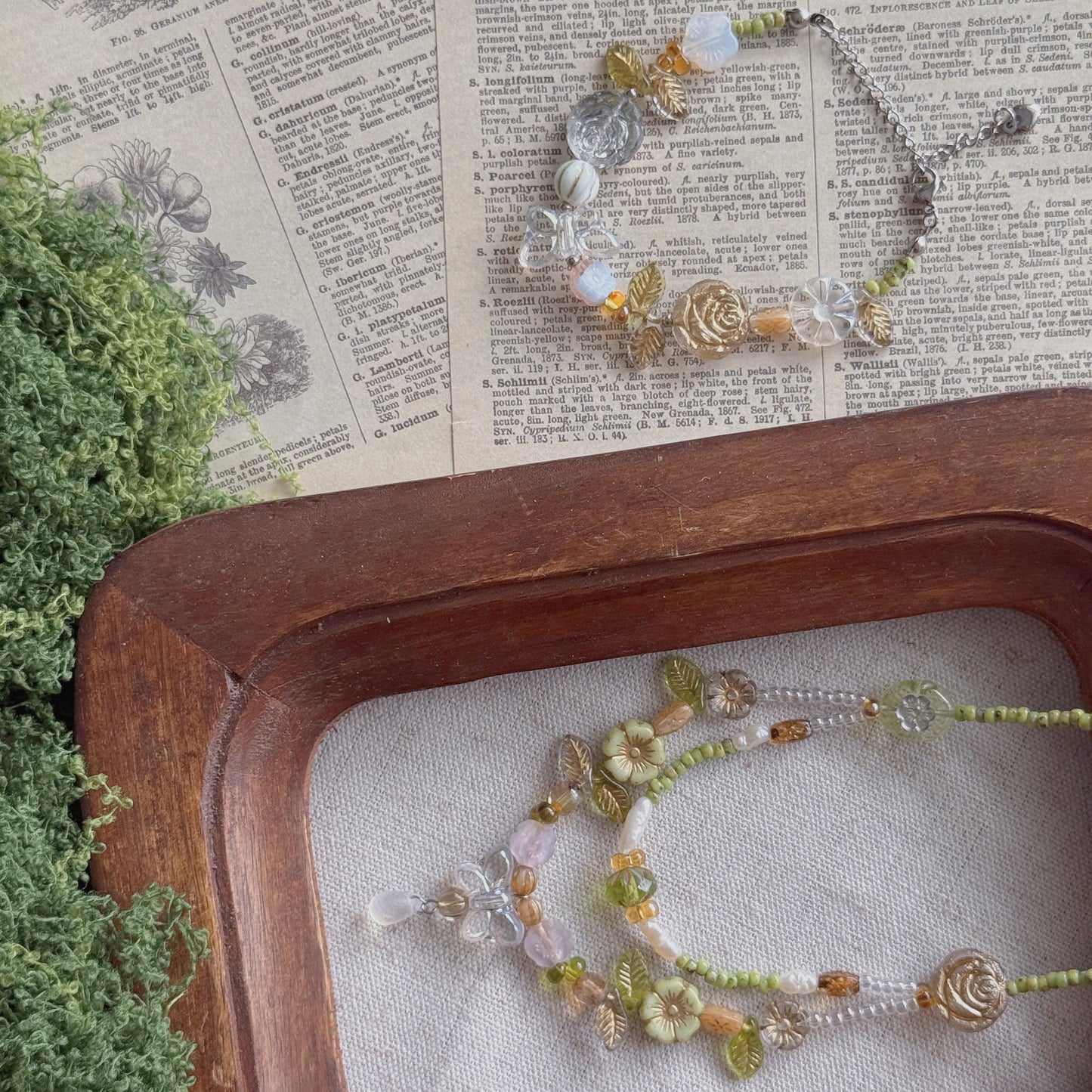 A delicate beaded jewelry set with green, amber, and clear Czech glass beads, displayed on a rustic wooden tray over vintage book pages with green foliage.