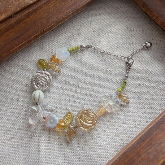 A delicate beaded bracelet with rose and leaf Czech glass charms, displayed on a textured beige fabric inside a rustic wooden frame.