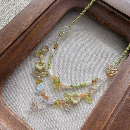 A delicate beaded necklace with green, yellow, and white Czech glass beads, floral charms, and a small hanging crystal pendant, displayed inside a rustic wooden frame on a linen surface.