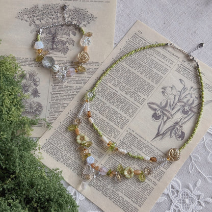 A beaded necklace and matching bracelet displayed on vintage book pages with a green plant sprig. The jewelry features light green beads, clear Czech glass beads, amber-colored beads, and a small gold rose charm.