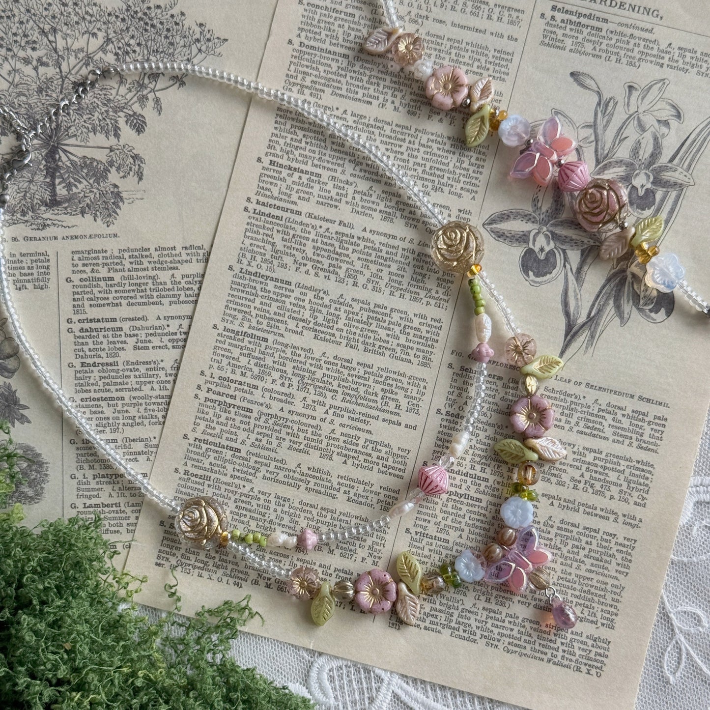 A delicate beaded necklace with pink and cream floral charms and green leaves Czech glass beads, styled on an antique book page with a sprig of greenery in the corner.