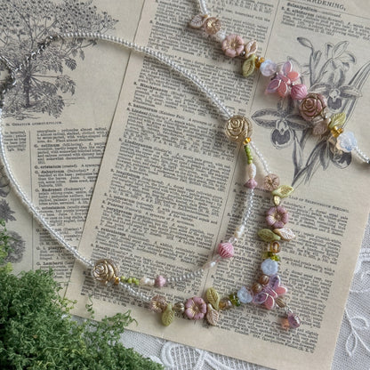 A delicate beaded necklace with pink and cream floral charms and green leaves Czech glass beads, styled on an antique book page with a sprig of greenery in the corner.