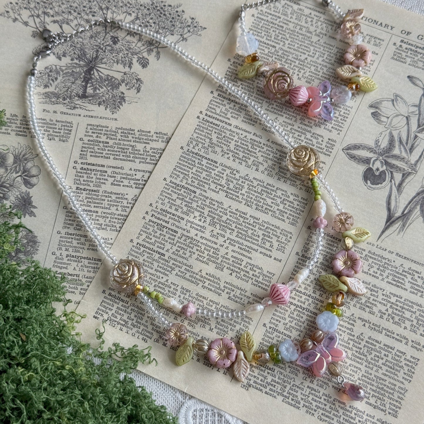 A delicate beaded necklace with floral and leaf charms, displayed on an open vintage book with green foliage in the foreground. The necklace features white pearl-like beads, pink and lavender flowers, green leaves, and small gold-toned accents.
