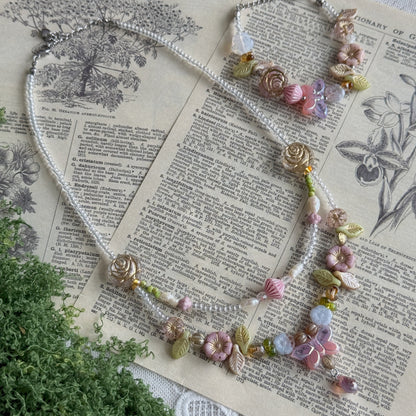A delicate beaded necklace with floral and leaf charms, displayed on an open vintage book with green foliage in the foreground. The necklace features white pearl-like beads, pink and lavender flowers, green leaves, and small gold-toned accents.