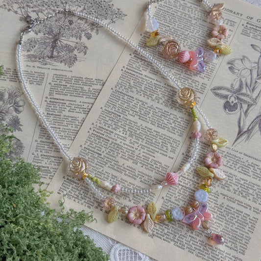 A delicate beaded necklace with floral and shell-like charms in pastel pink, peach, yellow, and lavender, displayed on vintage book pages with green foliage and lace as props.