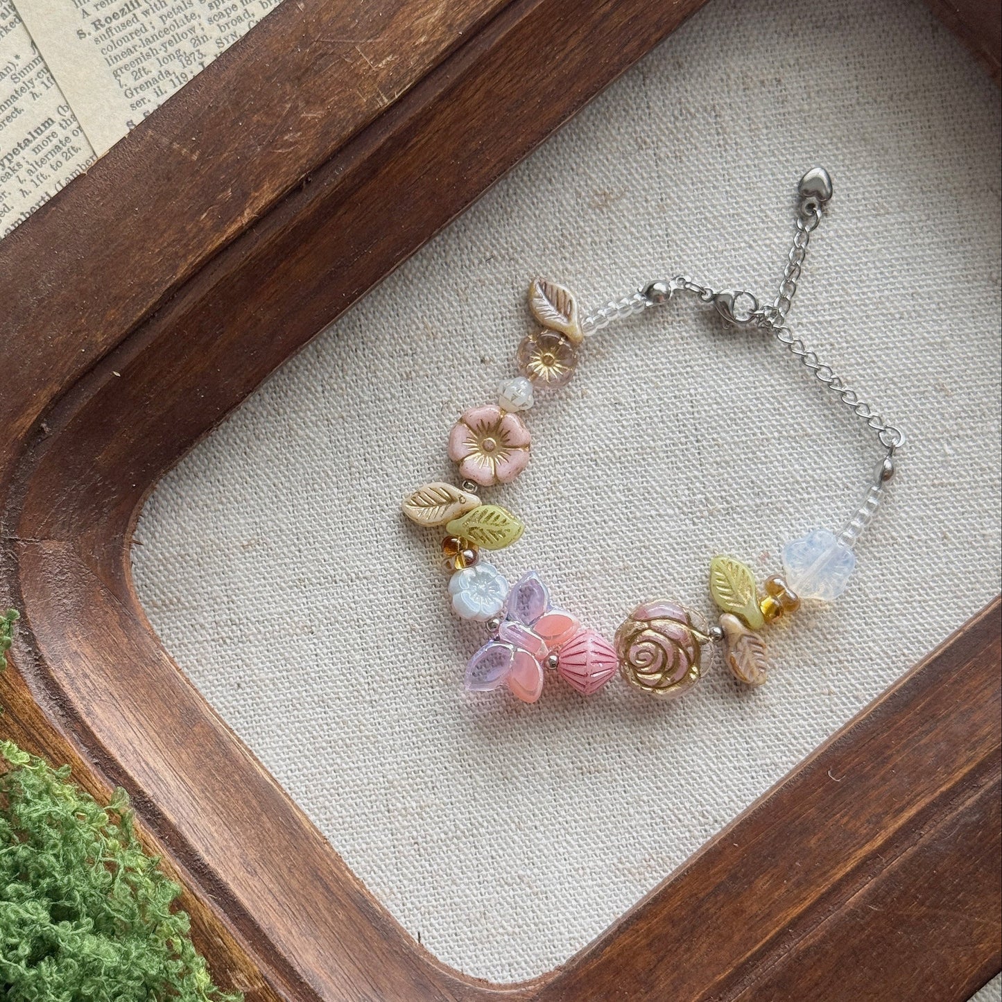 A delicate silver chain bracelet with pastel pink, lavender, and yellow floral Czech glass charms, displayed on a beige fabric surface inside a rustic wooden tray.
