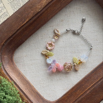 A delicate silver chain bracelet with pastel pink, lavender, and yellow floral Czech glass charms, displayed on a beige fabric surface inside a rustic wooden tray.