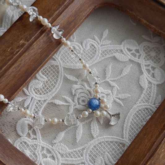 A delicate beaded necklace with white pearls, clear quartz, and a 5A-grade Kyanite bead, displayed on a white embroidered lace surface inside a wooden frame.