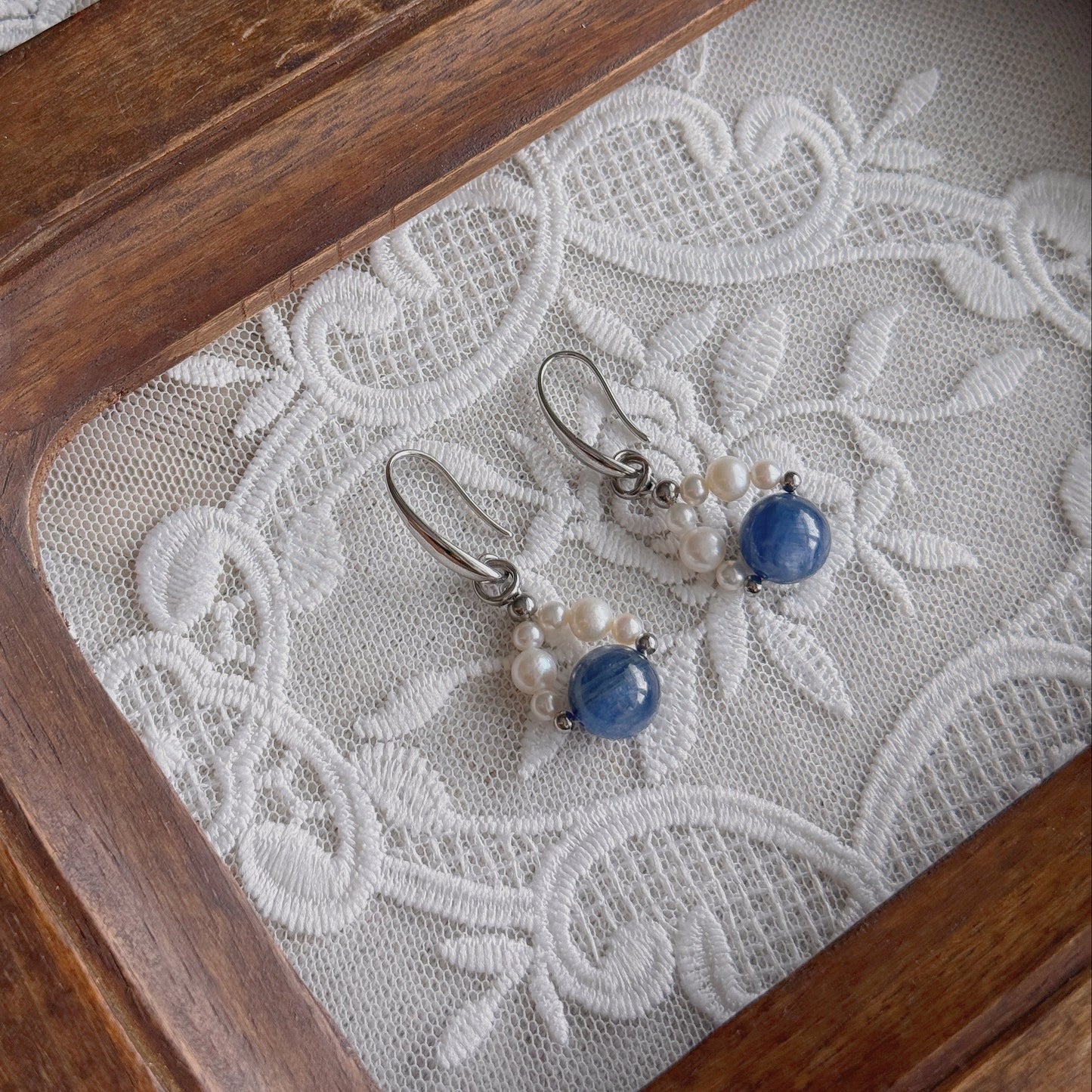 A pair of silver drop earrings with small freshwater pearls and round natural Kyanite beads, displayed on white embroidered lace inside a wooden tray.