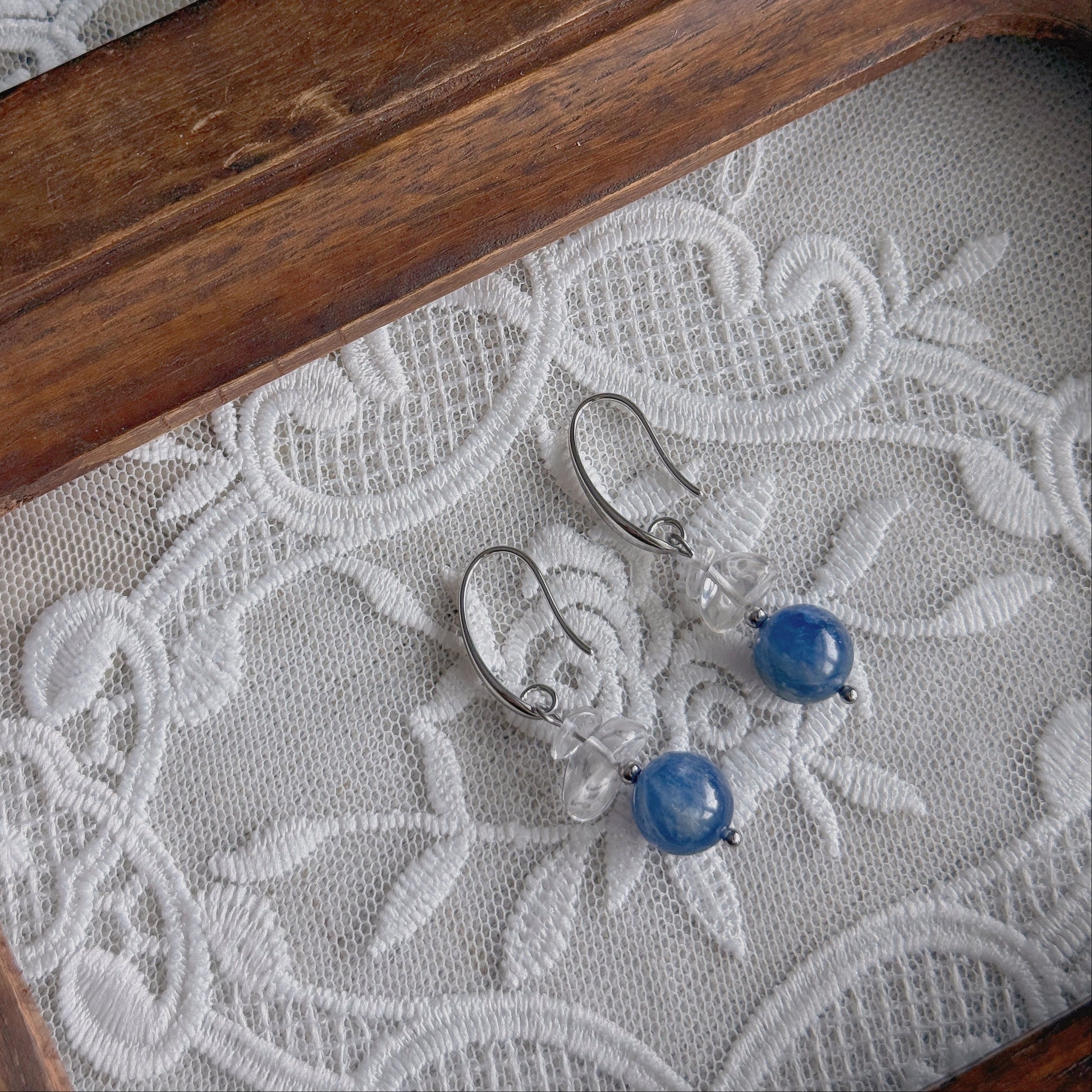 A pair of silver-toned drop earrings with clear quarts and round natural Kyanite beads, displayed on white embroidered lace inside a wooden tray.