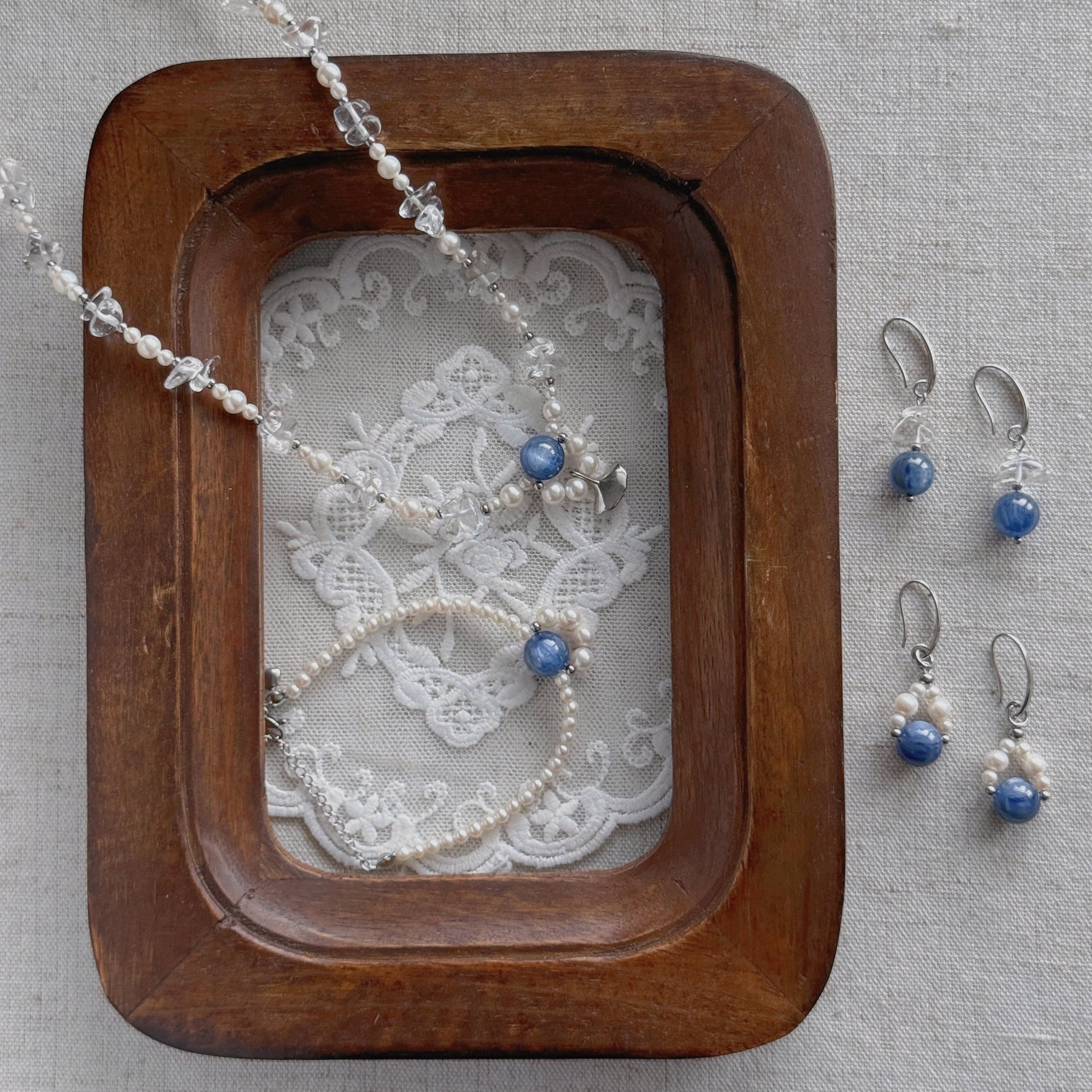 Silver-toned jewelry set featuring a beaded necklace, bracelet, and matching drop earrings with blue beads, arranged on a lace insert inside a wooden tray against a neutral fabric background.