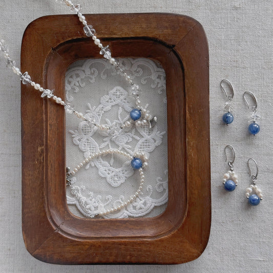 Silver-toned jewelry set featuring a beaded necklace, bracelet, and matching drop earrings with blue beads, arranged on a lace insert inside a wooden tray against a neutral fabric background.