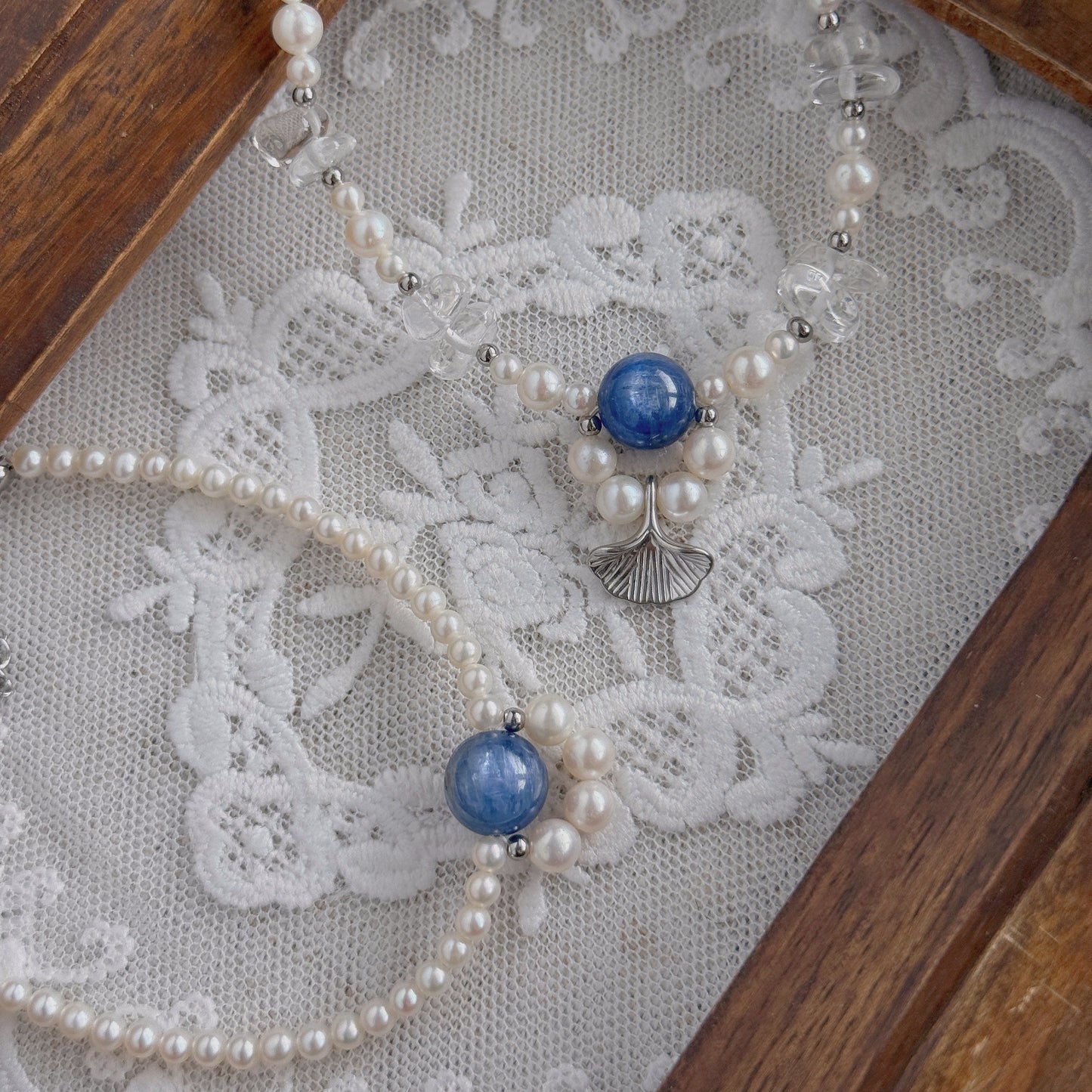 A delicate beaded necklace and a bracelet with freshwater pearls, clear quartz, and blue Kyanite beads, displayed on a white lace surface inside a wooden tray.