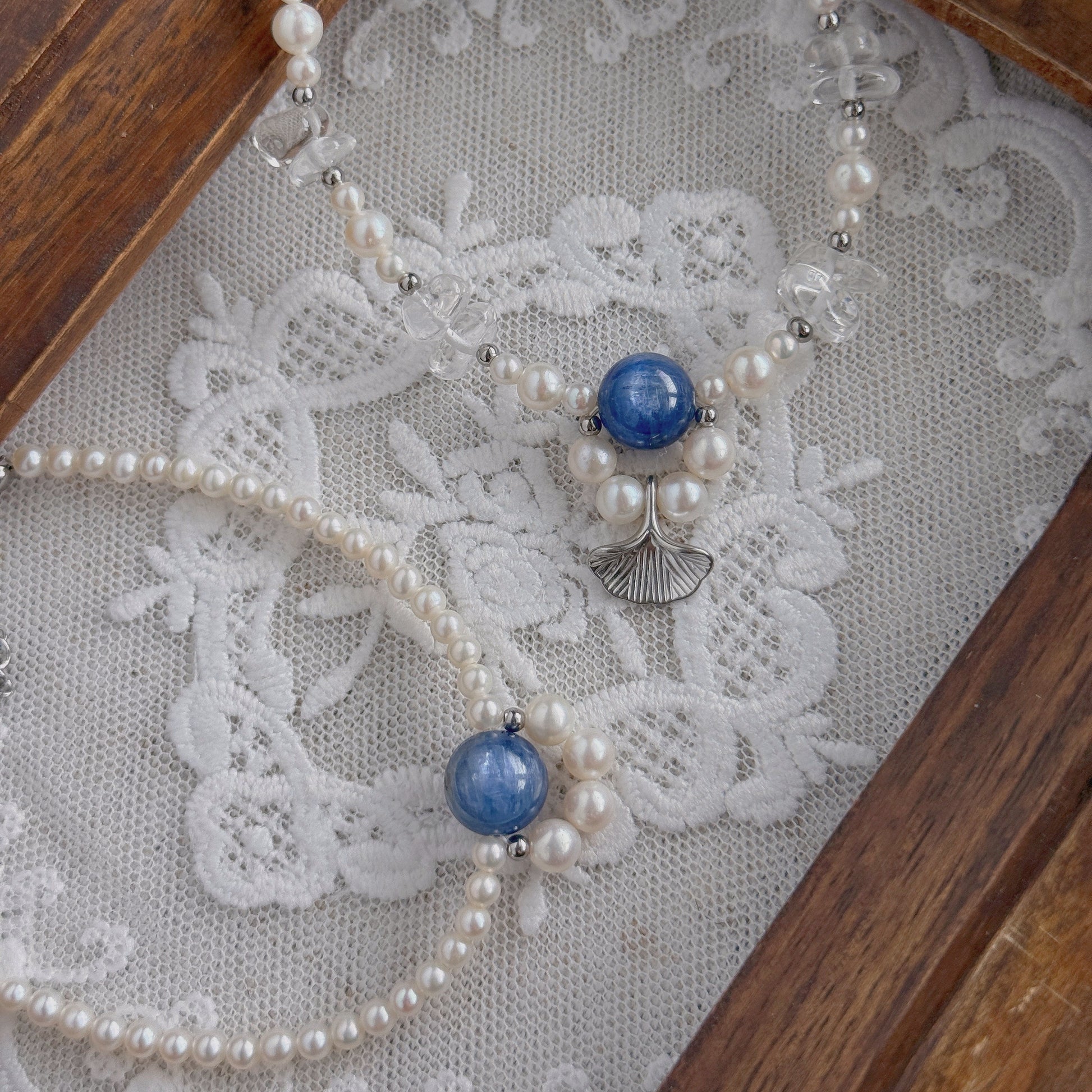 A delicate beaded necklace and a bracelet with freshwater pearls, clear quartz, and blue Kyanite beads, displayed on a white lace surface inside a wooden tray.
