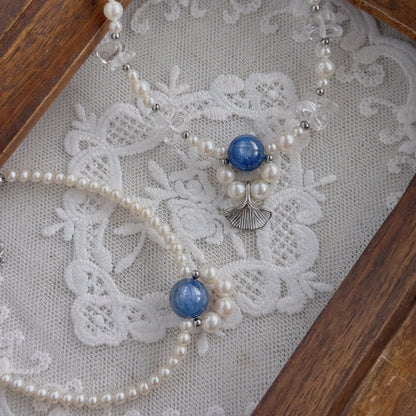 A delicate beaded necklace and a bracelet with freshwater pearls, clear quartz, and blue Kyanite beads, displayed on a white lace surface inside a wooden tray.
