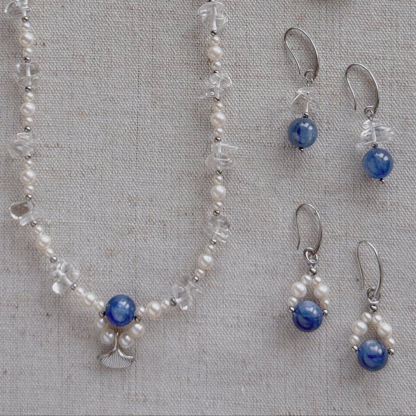 A jewelry set laid flat on a textured beige fabric background, including a long necklace and two matching drop earrings. The pieces feature freshwater pearls, clear quartz, silver-toned metal findings, and round natural Kyanite stones.