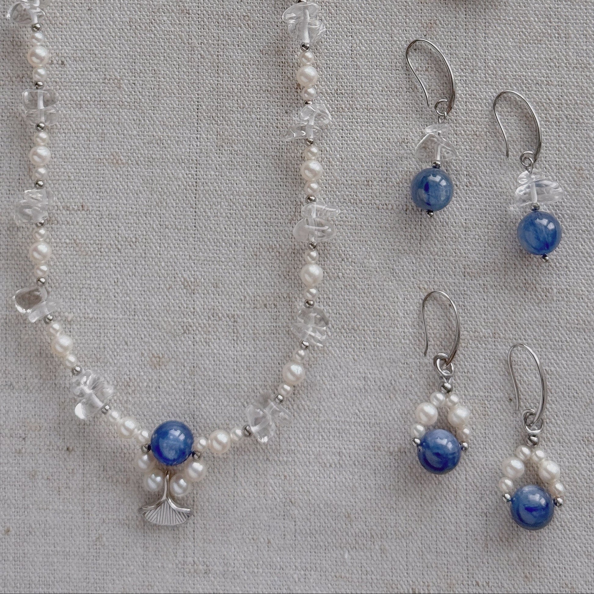 A jewelry set laid flat on a textured beige fabric background, including a long necklace and two matching drop earrings. The pieces feature freshwater pearls, clear quartz, silver-toned metal findings, and round natural Kyanite stones.