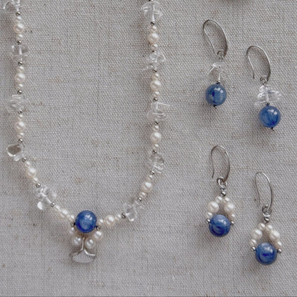 A jewelry set laid flat on a textured beige fabric background, including a long necklace and two matching drop earrings. The pieces feature freshwater pearls, clear quartz, silver-toned metal findings, and round natural Kyanite stones.