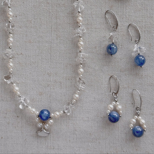 A jewelry set laid flat on a textured beige fabric background, including a long necklace and two matching drop earrings. The pieces feature freshwater pearls, clear quartz, silver-toned metal findings, and round natural Kyanite stones.