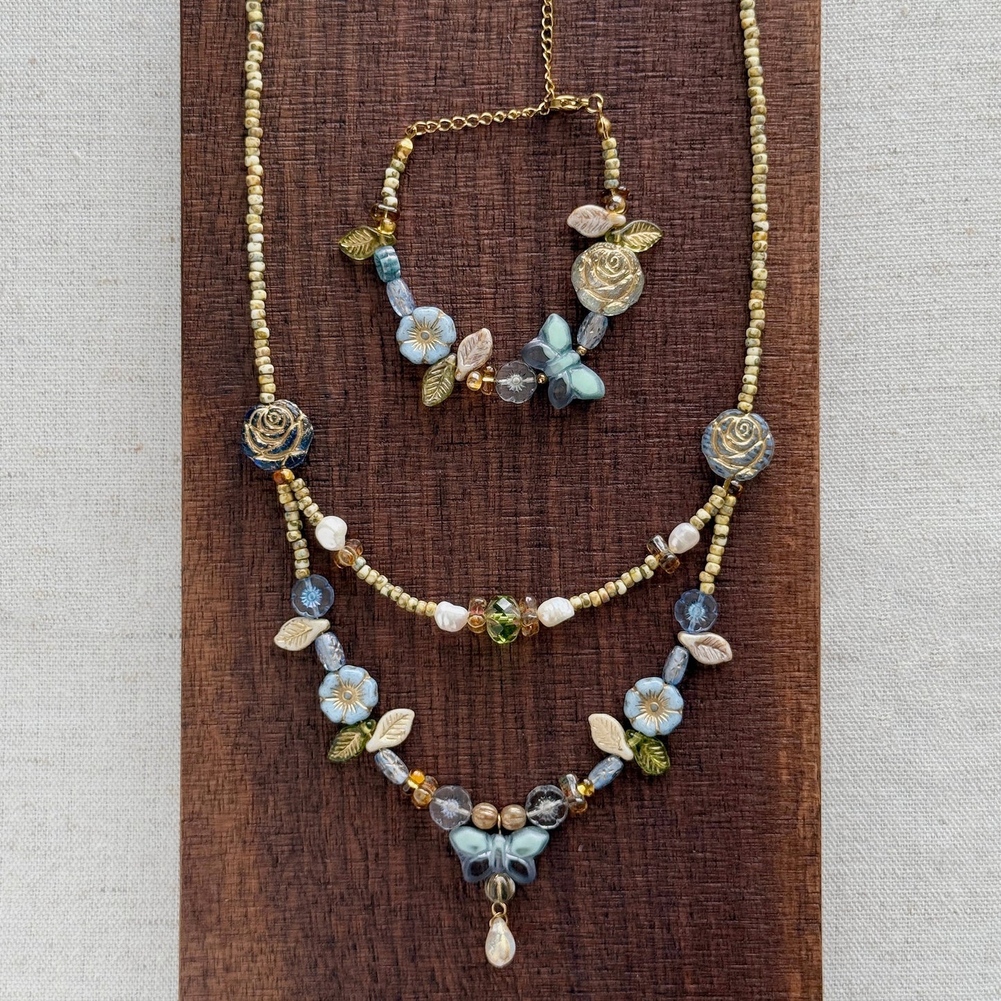 A bohemian-style jewelry set featuring a gold-tone chain necklace and matching bracelet adorned with blue, white, and green floral beads, displayed on a dark wood block against a neutral linen background.