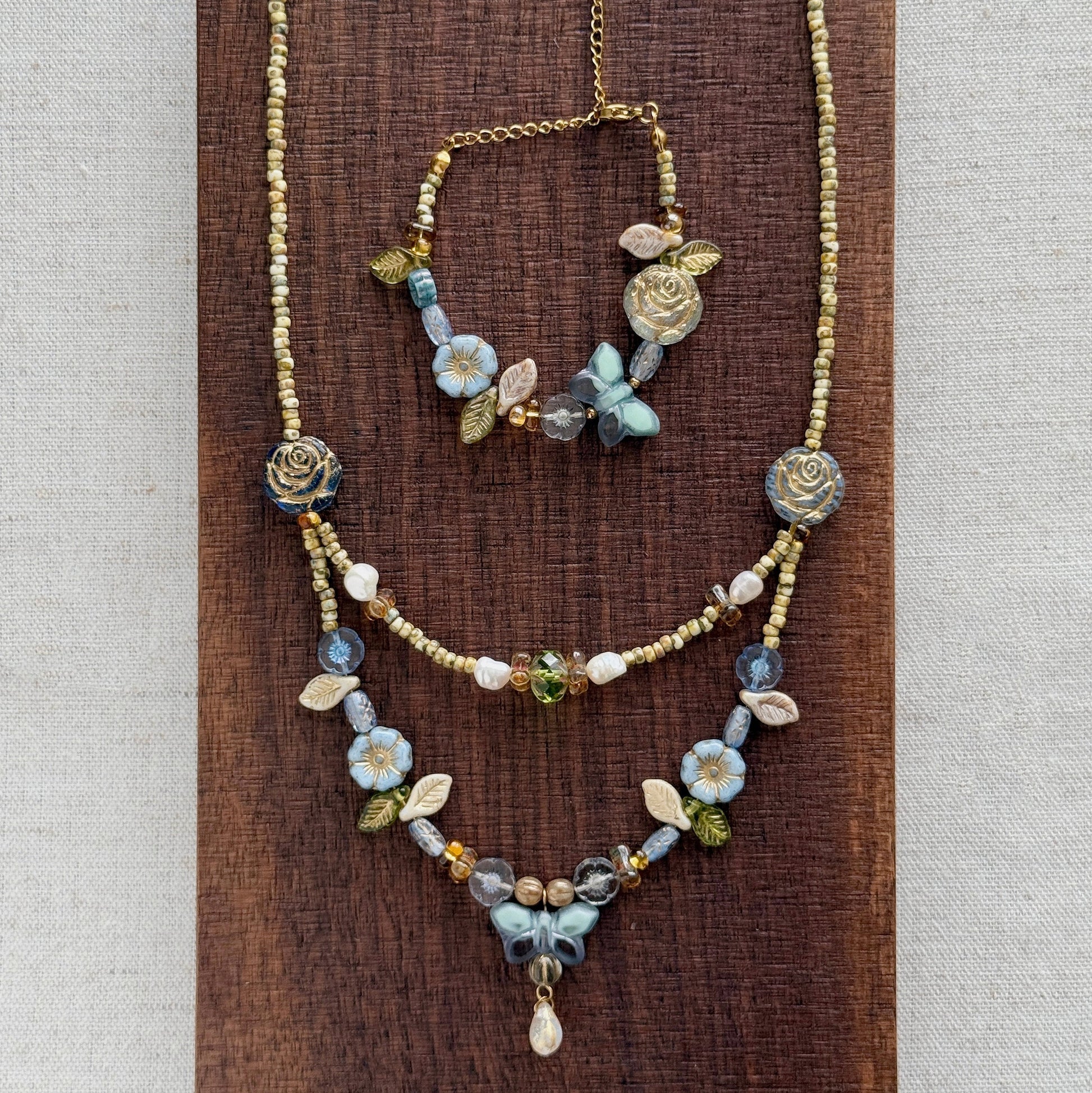 A bohemian-style jewelry set featuring a gold-tone chain necklace and matching bracelet adorned with blue, white, and green floral beads, displayed on a dark wood block against a neutral linen background.