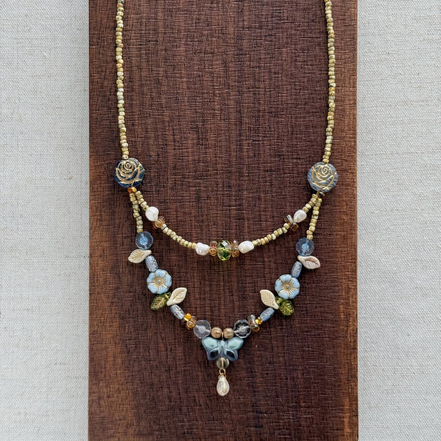 A delicate layered necklace displayed on a dark brown textured wooden block against a light beige linen background. The necklace features a gold-tone chain with blue and white floral bead details, a short beaded section, and a longer floral pendant section with a small teardrop charm.