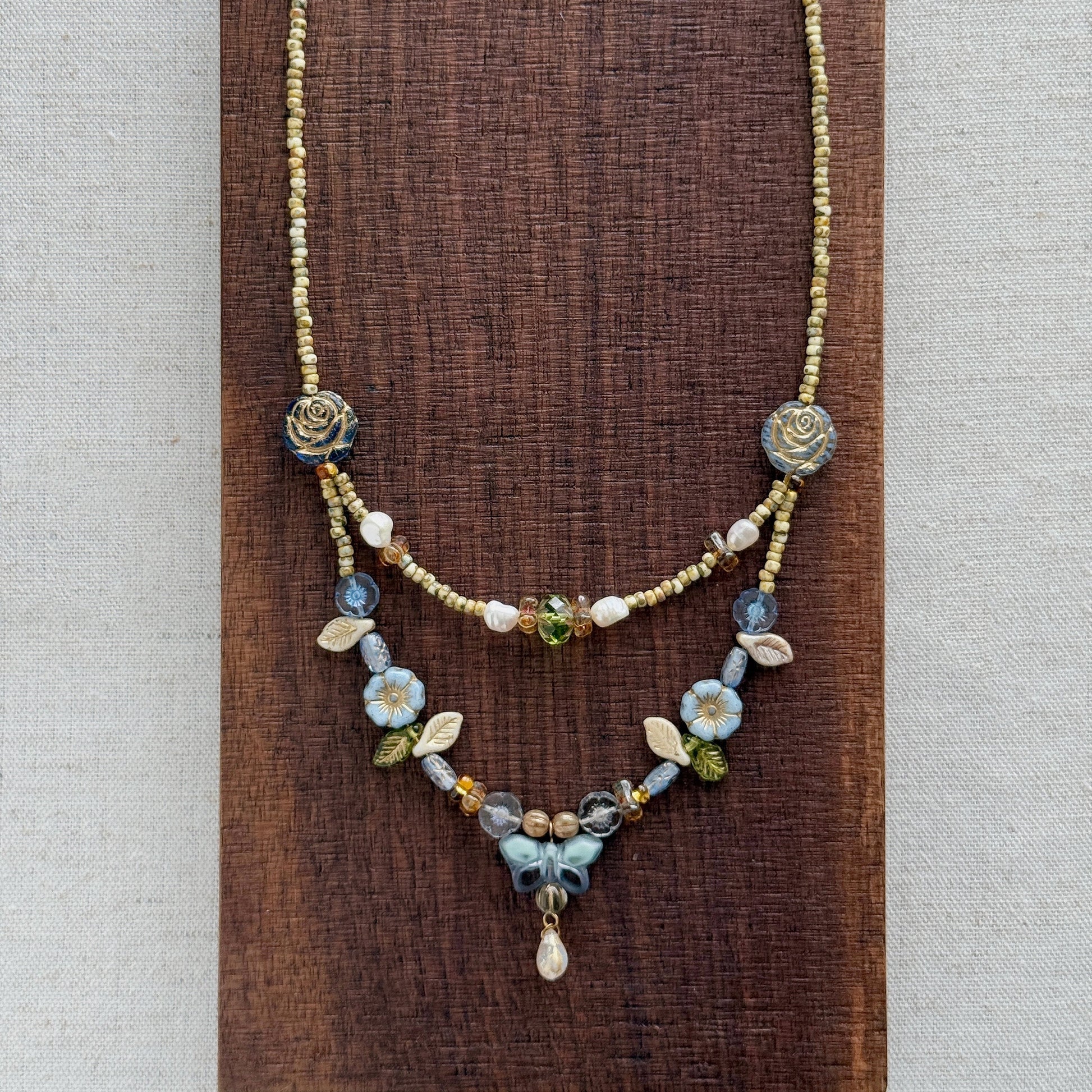 A delicate layered necklace displayed on a dark brown textured wooden block against a light beige linen background. The necklace features a gold-tone chain with blue and white floral bead details, a short beaded section, and a longer floral pendant section with a small teardrop charm.