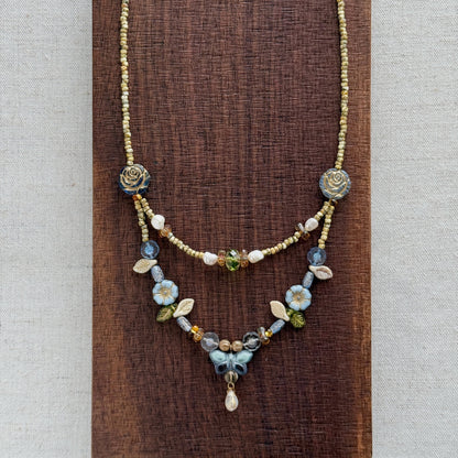 A delicate layered necklace displayed on a dark brown textured wooden block against a light beige linen background. The necklace features a gold-tone chain with blue and white floral bead details, a short beaded section, and a longer floral pendant section with a small teardrop charm.