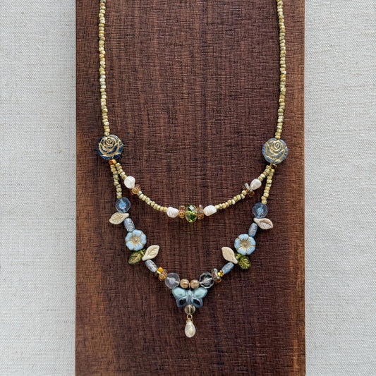 A delicate layered necklace displayed on a dark brown textured wooden block against a light beige linen background. The necklace features a gold-tone chain with blue and white floral bead details, a short beaded section, and a longer floral pendant section with a small teardrop charm.