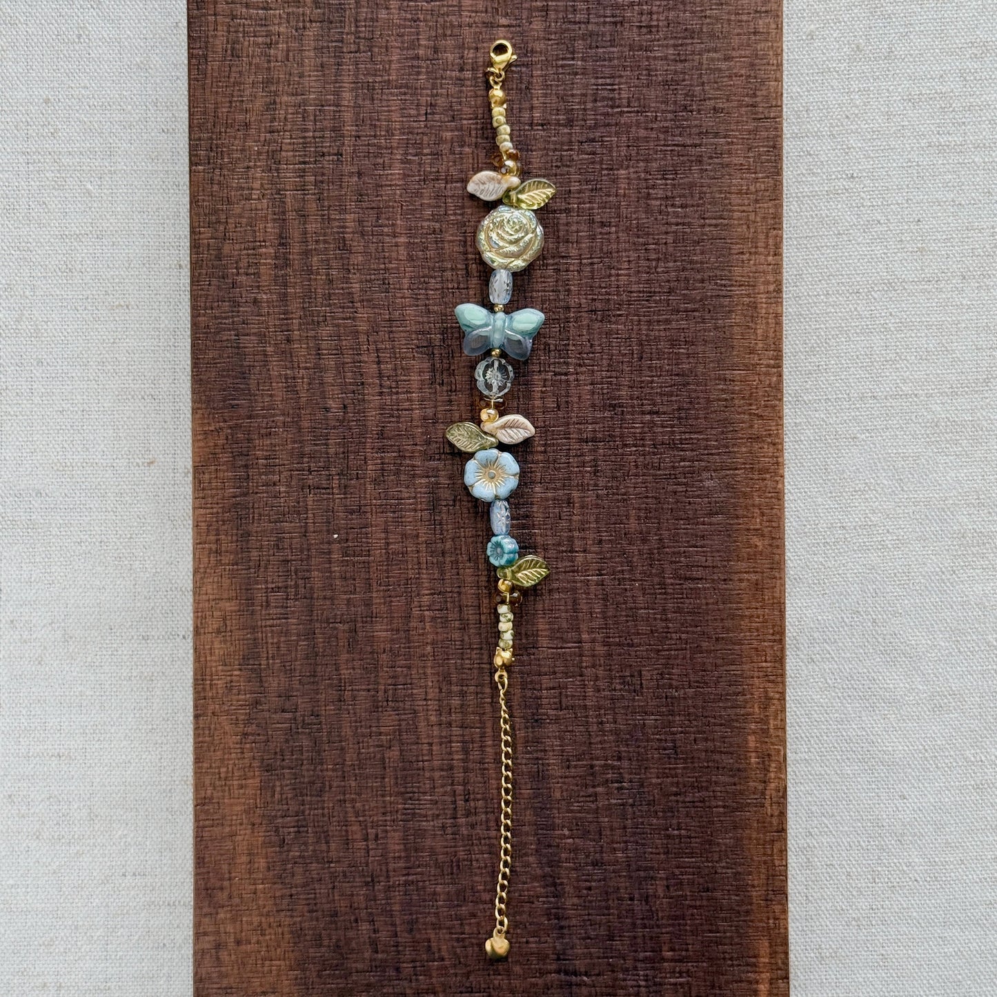A delicate beaded bracelet with gold-toned metal beads, light blue floral elements, and a small butterfly charm, displayed vertically on a dark brown wooden block against a light beige textured fabric background.