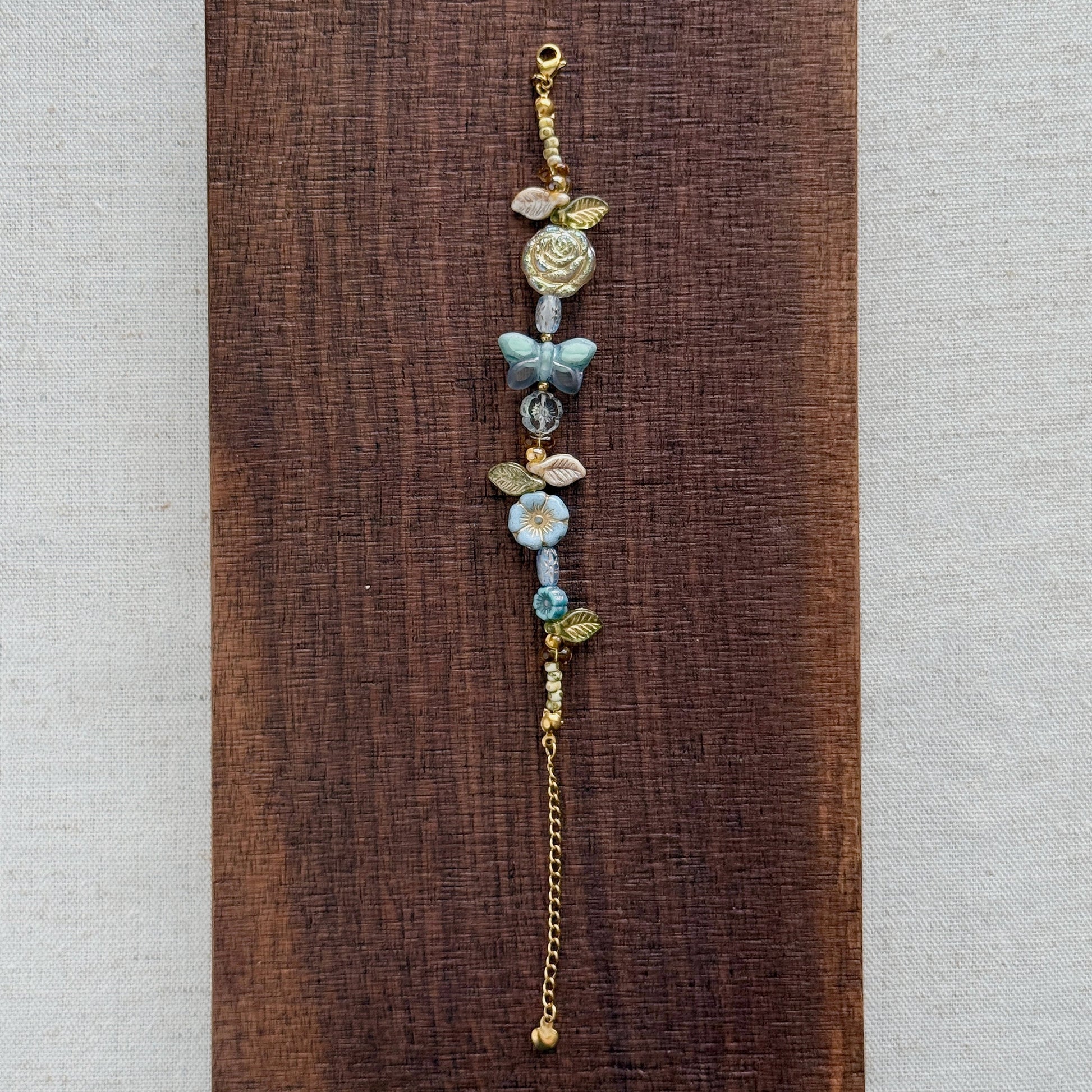 A delicate beaded bracelet with gold-toned metal beads, light blue floral elements, and a small butterfly charm, displayed vertically on a dark brown wooden block against a light beige textured fabric background.