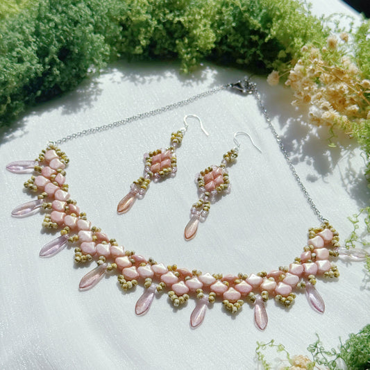Pink beaded statement necklace with matching dangle earrings, featuring floral petal design and soft romantic tones, handmade jewelry on a textured fabric background