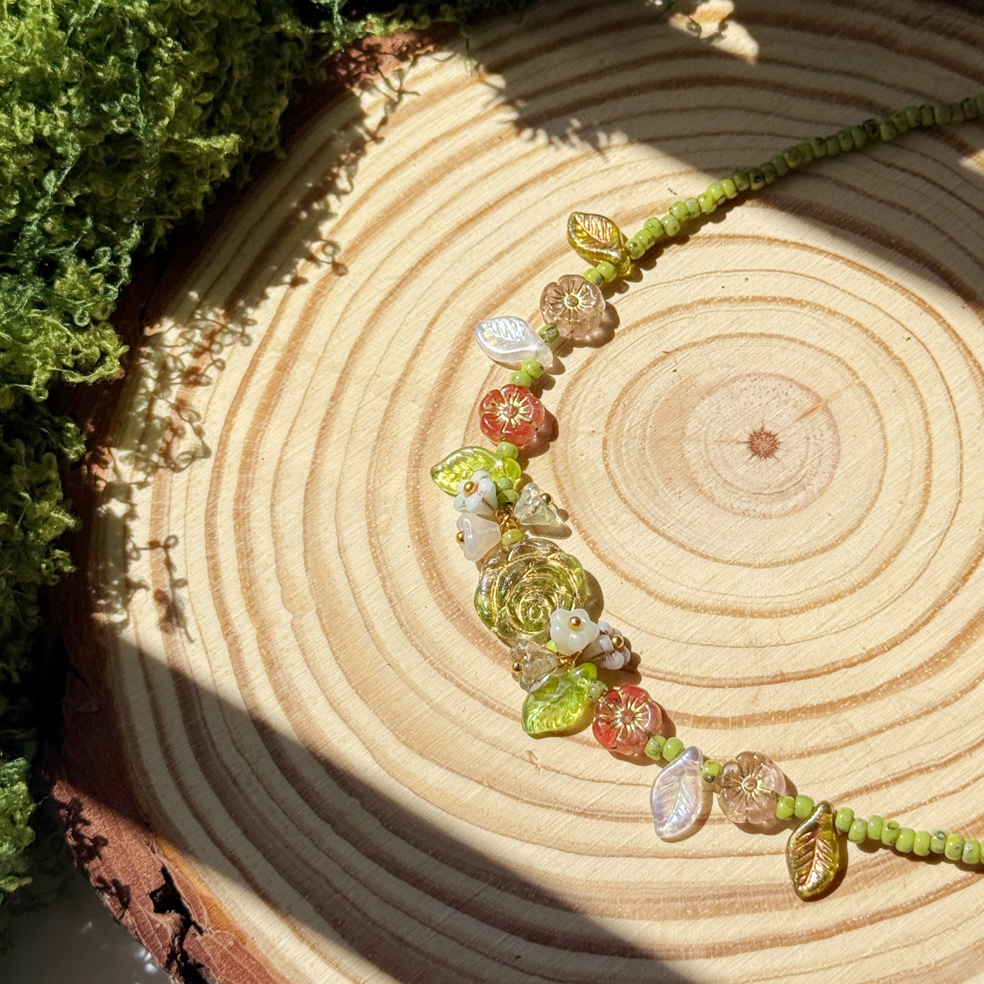 Necklace with green leaf beads and red flower beads on a wooden surface with moss