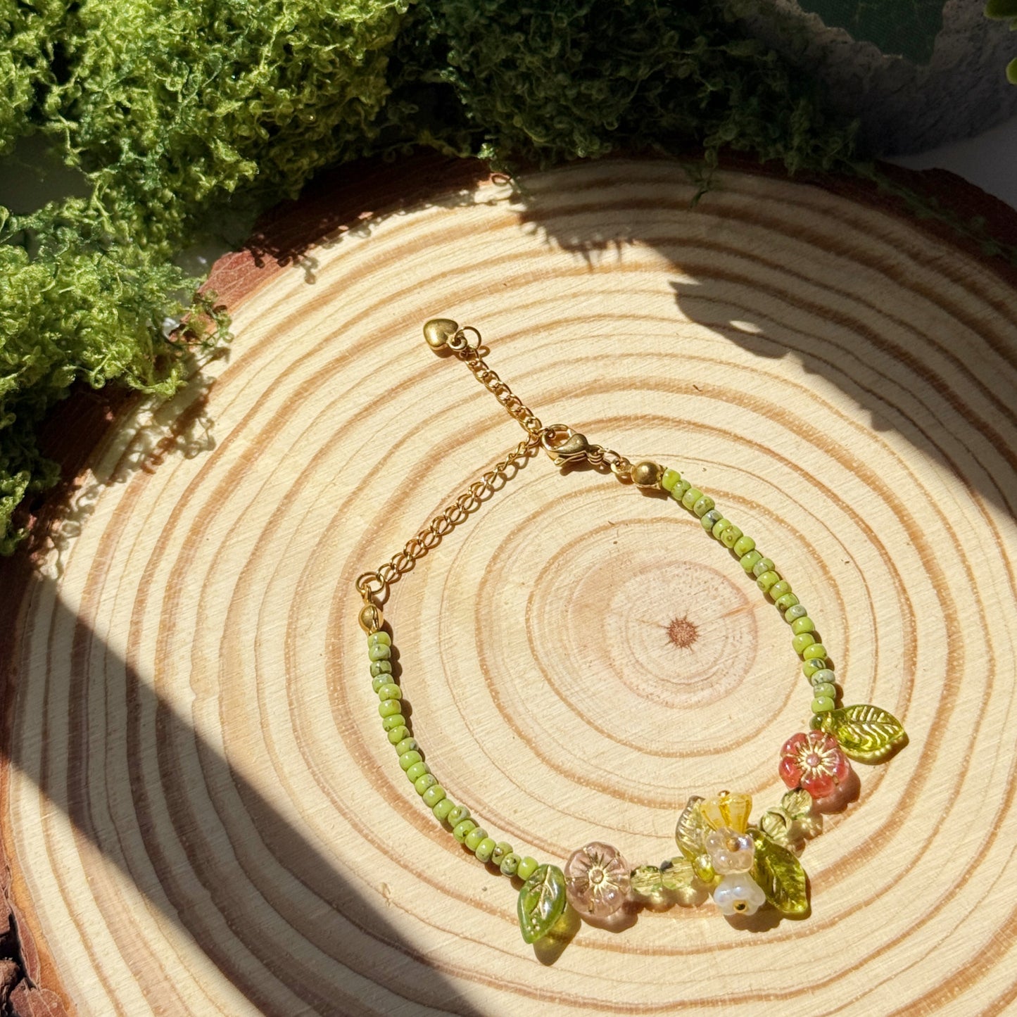 Bracelet with green leaf beads and floral charms on a wooden surface with greenery
