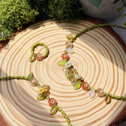 Beaded necklace, bracelet, and ring on a wooden surface with greenery and flowers in the background
