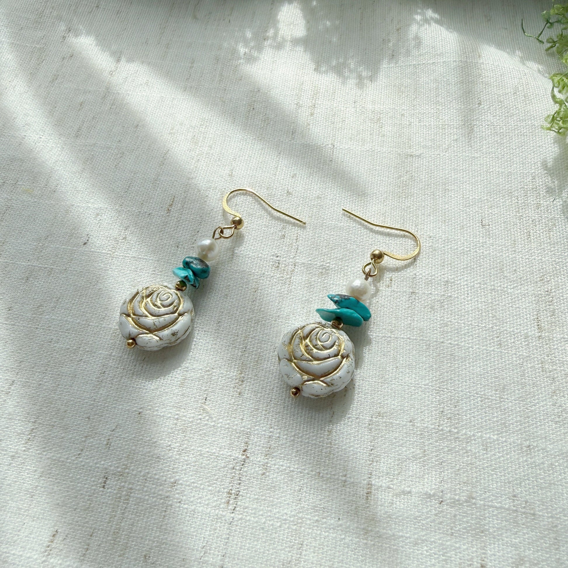 Handmade earrings featuring natural turquoise, freshwater pearls, and Czech glass rose beads displayed on a soft linen background.