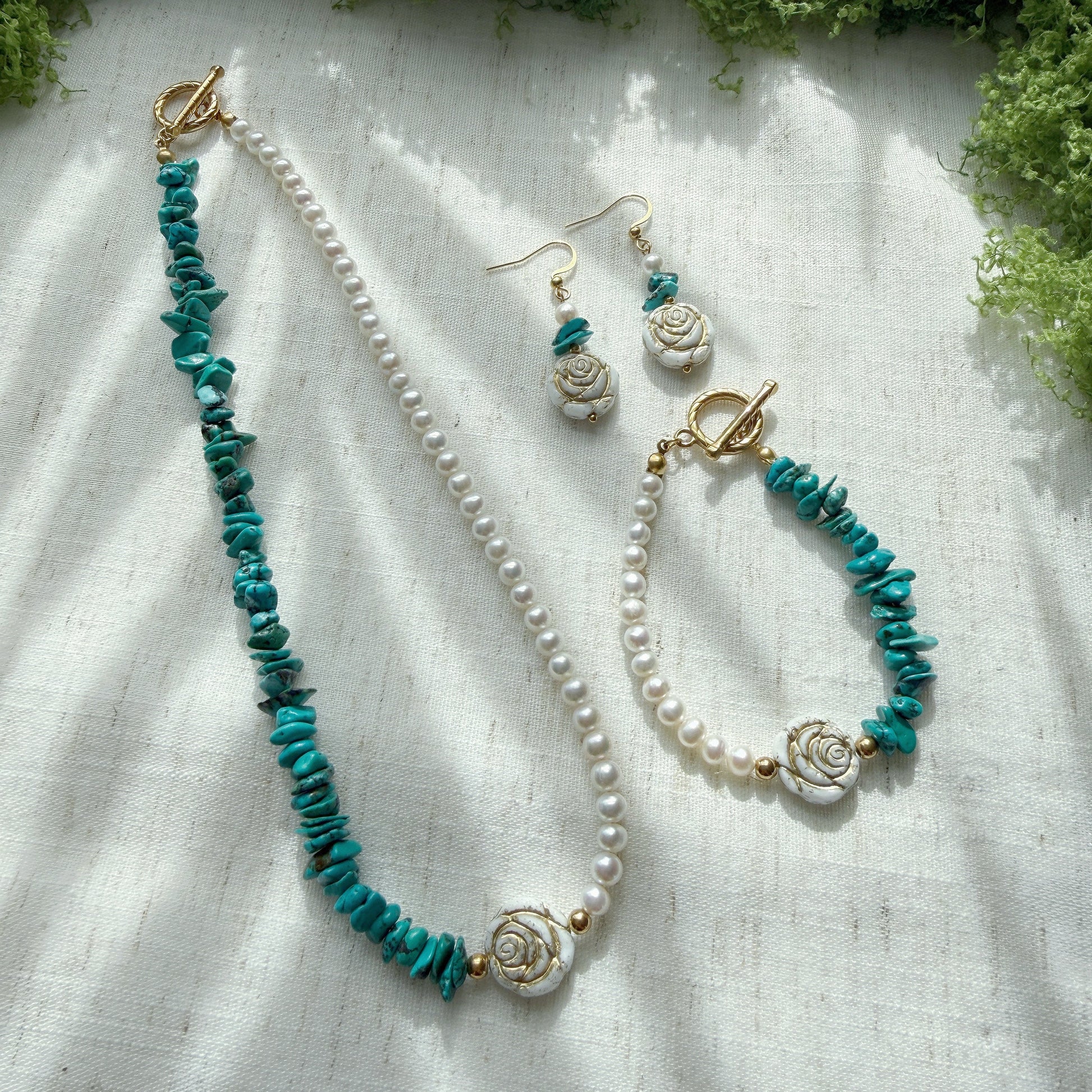 Handmade jewelry set featuring natural turquoise, freshwater pearls, and Czech glass rose beads, including a necklace, bracelet, and matching earrings displayed on a soft linen background.