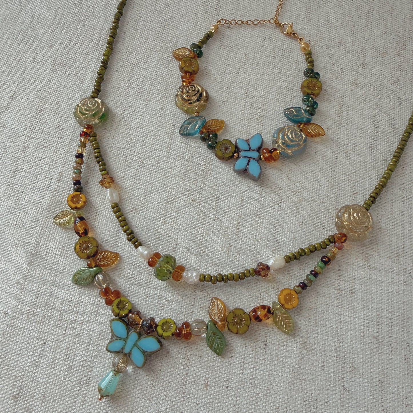 A jewelry set featuring a double-strand necklace and a bracelet with olive green beaded chains, amber and turquoise glass beads, and two light blue butterfly charms, displayed on a textured beige linen background.