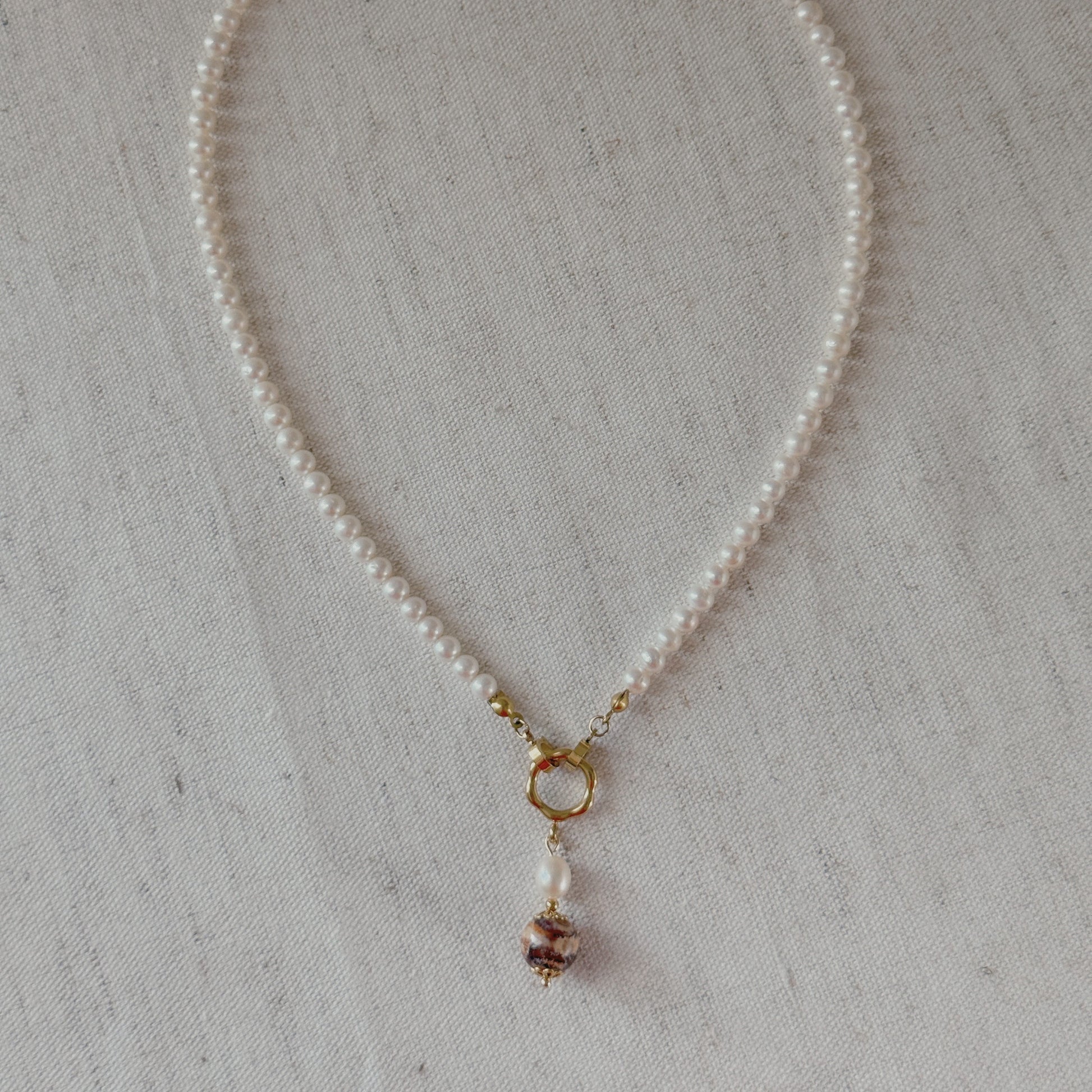 A delicate pearl necklace with a gold clasp and a agate dangling bead charm, displayed on a textured beige fabric background.