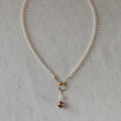 A delicate pearl necklace with a gold clasp and a agate dangling bead charm, displayed on a textured beige fabric background.
