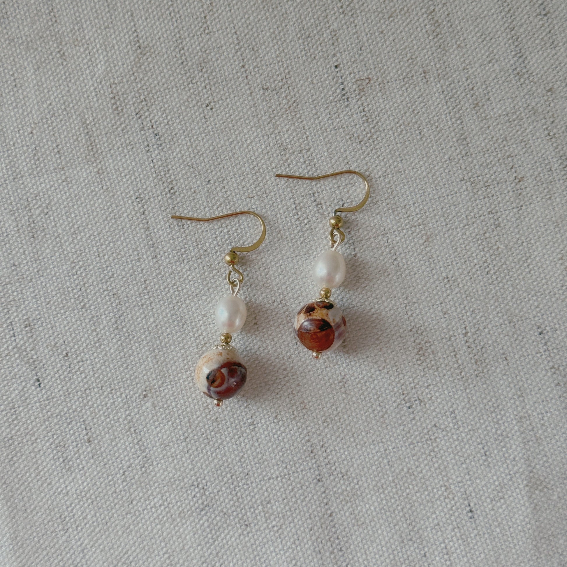 A pair of handmade dangle earrings featuring white freshwater pearl beads and agate beads on gold-tone hooks, displayed on a textured off-white linen background.