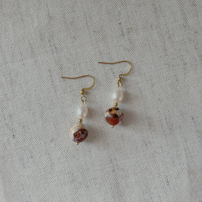 A pair of handmade dangle earrings featuring white freshwater pearl beads and agate beads on gold-tone hooks, displayed on a textured off-white linen background.
