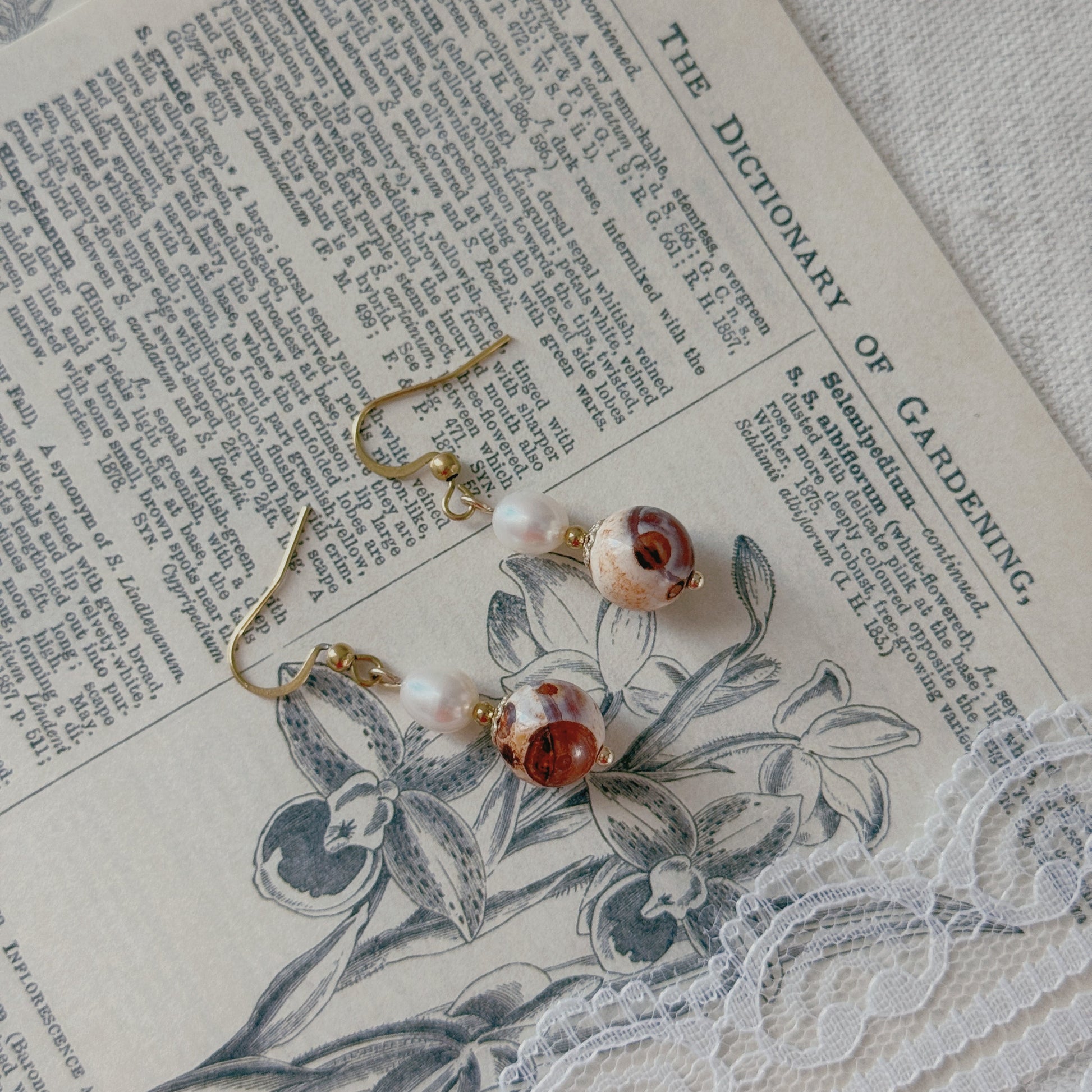 A pair of gold-tone dangle earrings with white freshwater pearl beads and agate beads, displayed on vintage dictionary pages with a floral print and lace.