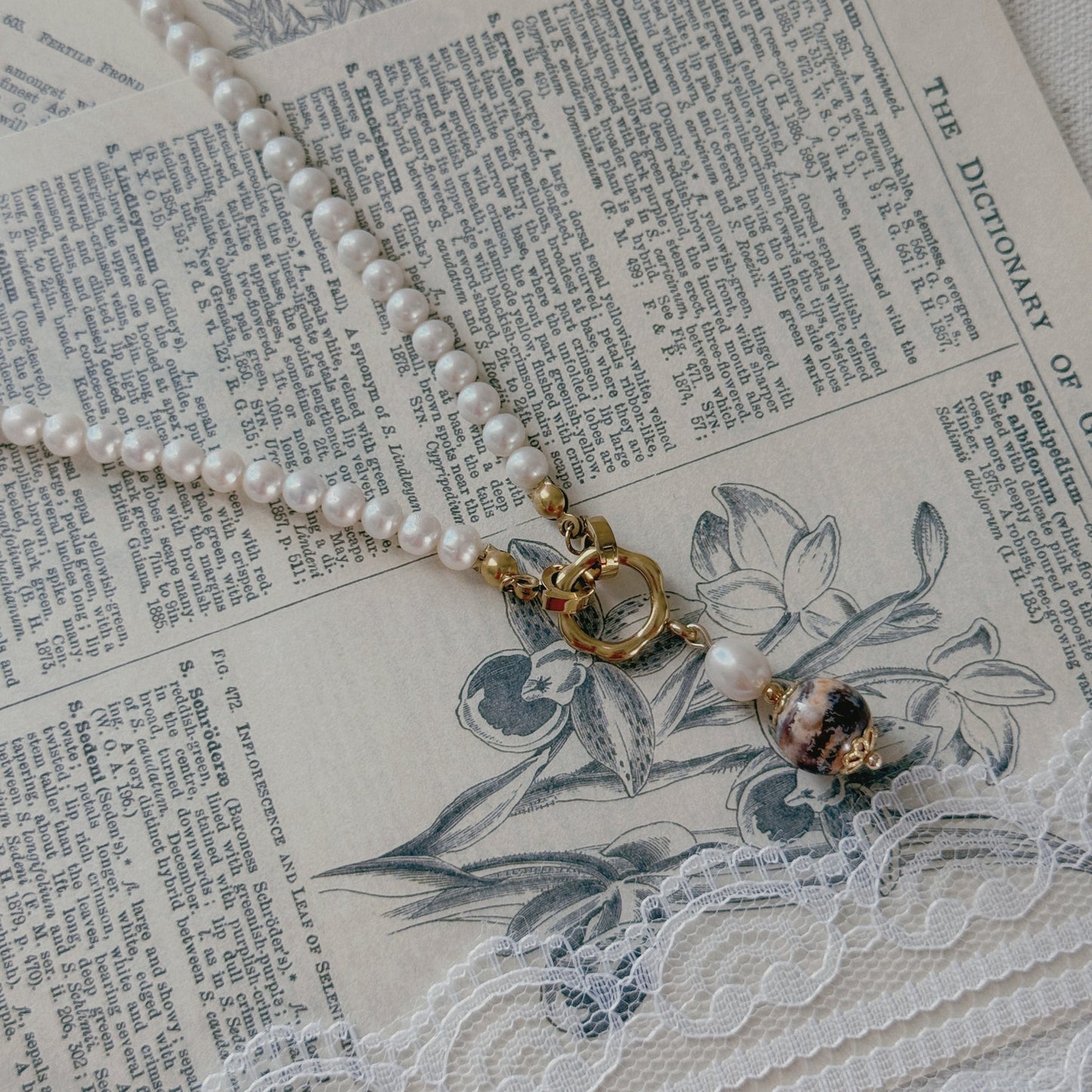 A pearl necklace with a gold clasp and a freshwater pearl and agate bead pendant, styled on vintage book pages with white lace.