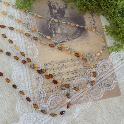 A beaded necklace with amber-brown, gold, and green beads draped over a vintage sheet music card, lace, and green foliage on a linen surface.