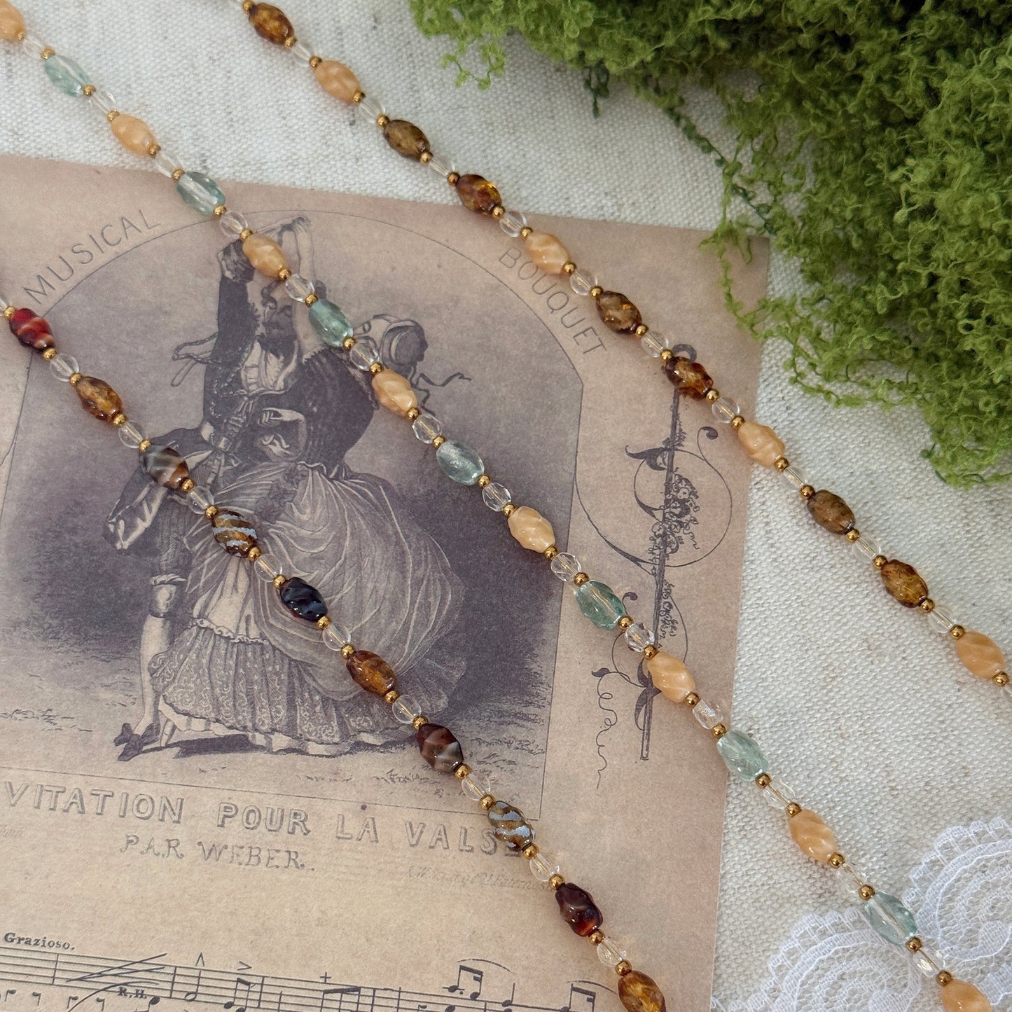 A beaded strand necklace or bracelet with faceted oval beads in amber, brown, pale green, and deep burgundy, threaded on a gold-tone chain. It is styled on a vintage sheet music print over a white lace cloth with green moss in the corner.