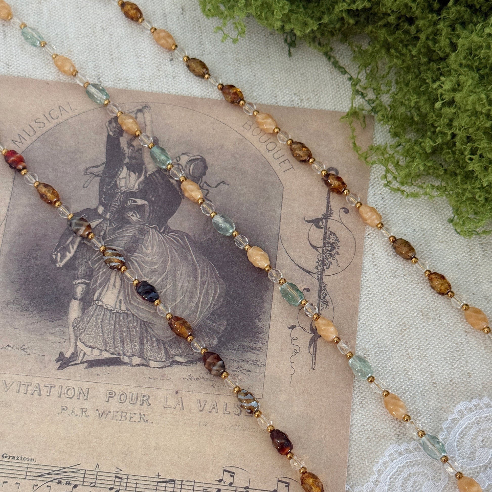 A beaded strand necklace or bracelet with faceted oval beads in amber, brown, pale green, and deep burgundy, threaded on a gold-tone chain. It is styled on a vintage sheet music print over a white lace cloth with green moss in the corner.