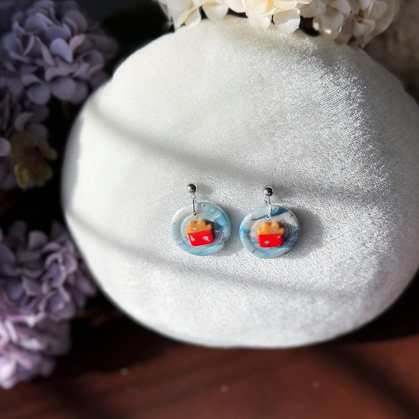 Earrings with a design of a small boat and red object on a white cushion, surrounded by flowers.