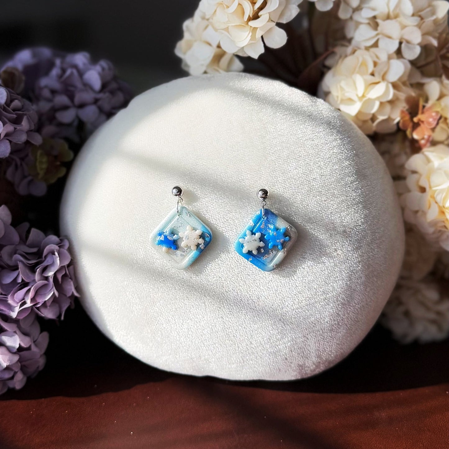 Pair of blue square earrings with Snowflake details on a white cushion surrounded by flowers.
